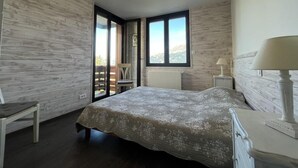 Un dormitorio con cama, una silla, una mesita de noche y una lámpara. Hay ventanas y vistas al exterior.