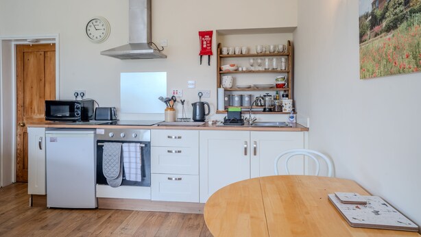 Une cuisine compacte équipée d’un four à micro-ondes, d’un grille-pain, d’une bouilloire et d’un petit réfrigérateur. On y trouve une table et une chaise en bois, ainsi qu’une horloge au mur.