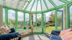 Un invernadero con estructura verde, ventanas amplias y vistas a un jardín. En el interior, hay un sofá y una pequeña mesa con una cesta.
