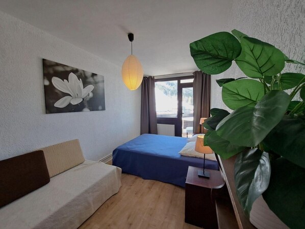 A bedroom with a bed, a sofa, a large plant, a painting, and a window with curtains.