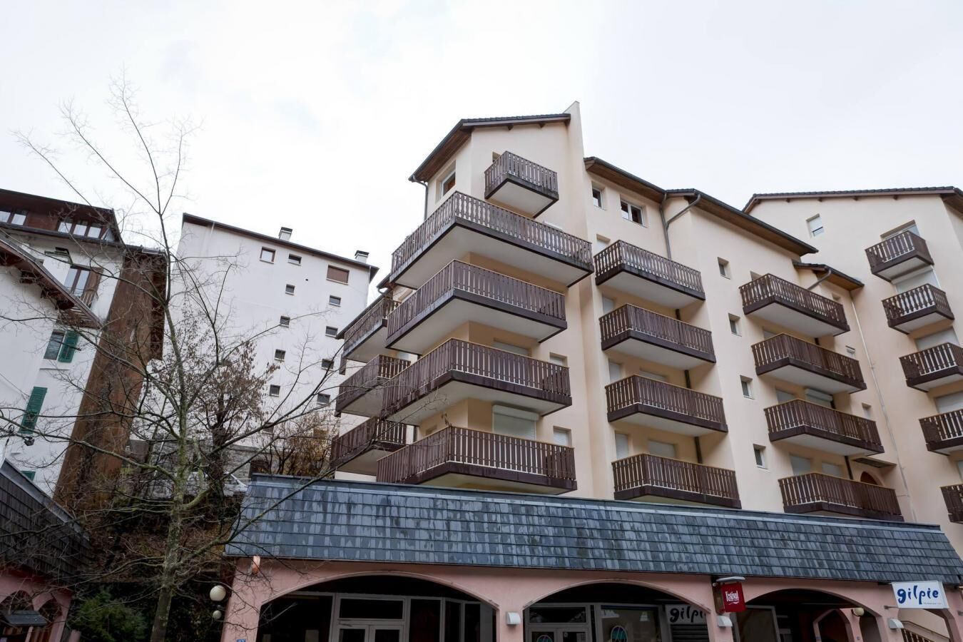 A multi-story residential building with balconies, a storefront with a sign, and a tree in the foreground.