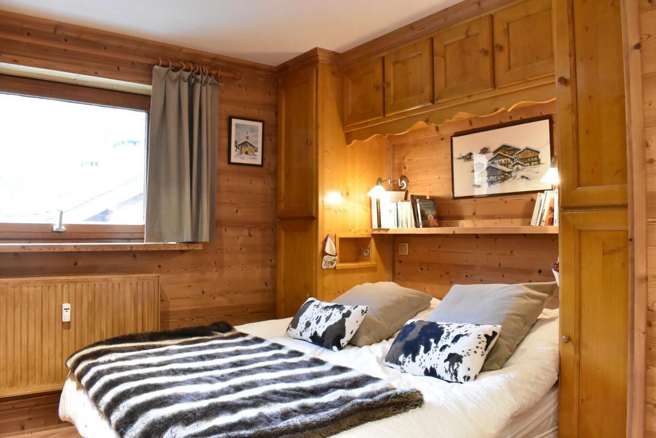 A cozy wooden room with a bed, a window with curtains, a shelf with books, and a wall-mounted light.