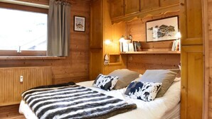 Una habitación acogedora de madera con una cama, un ventanal con cortinas, una estantería con libros y una lámpara fijada a la pared.