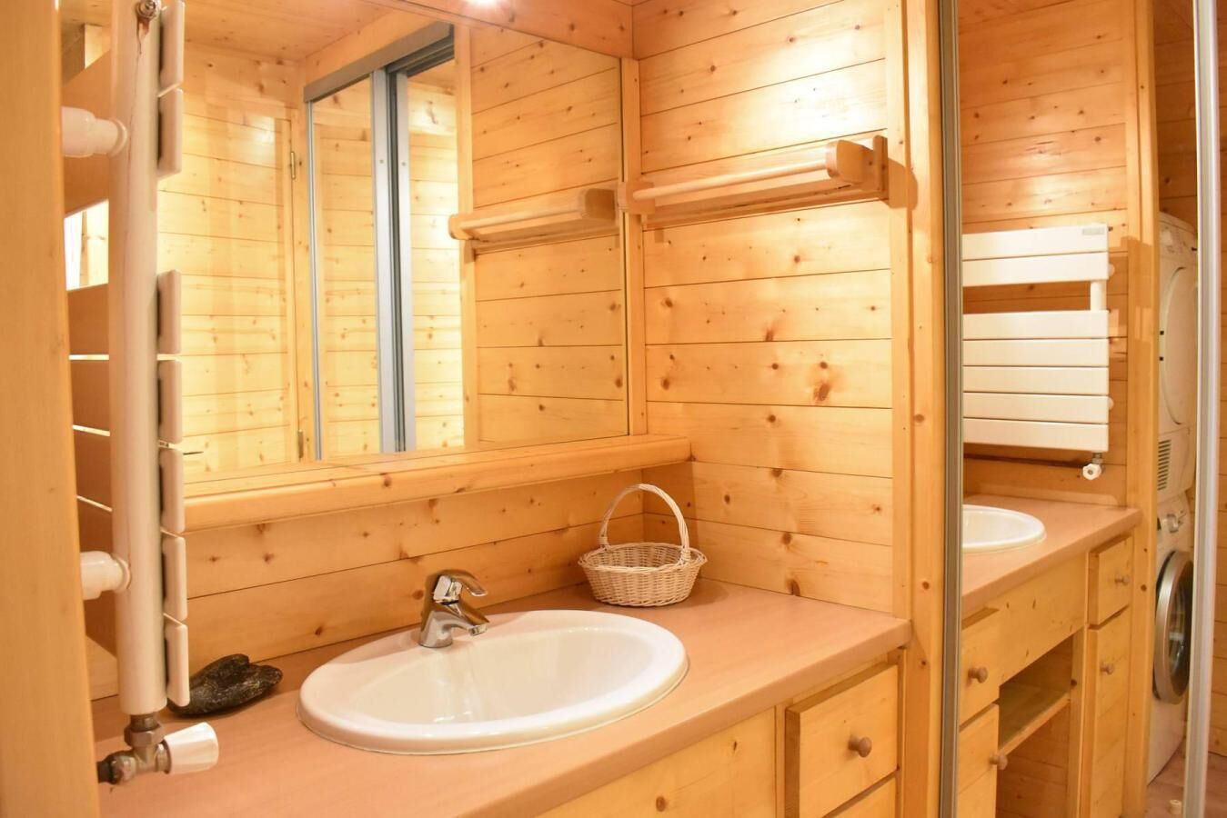 A wooden cabin bathroom with a large mirror, a sink, a washing machine, and a small basket.