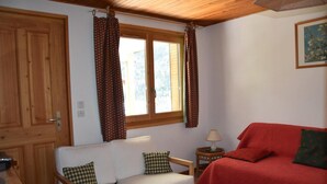A cozy room with a wooden ceiling, a white sofa with checkered pillows, a red sofa with a green pillow, a small wooden table with a lamp, and a window with patterned curtains.