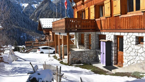 A wooden chalet with stone walls, a balcony, and a car parked in front.