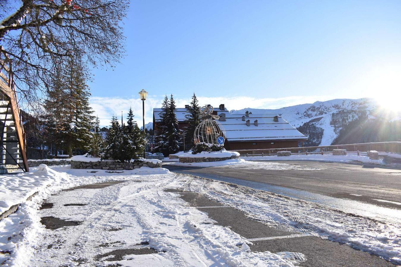 A snow-covered road leading to a building with a snow globe sculpture, surrounded by trees and mountains.