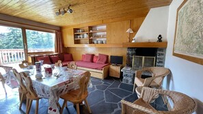 Quarto aconchegante com uma mesa de jantar posta para quatro pessoas, um sofá, cadeiras de vime, lareira e prateleiras de madeira com louças.