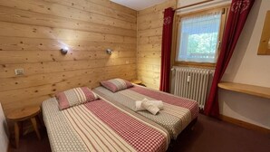 A room with two beds, wooden walls, a small stool, a window with red curtains, and a radiator.