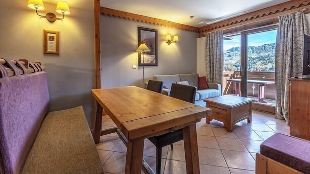 Habitación acogedora con una mesa de madera, un sofá y vistas a un paisaje montañoso a través de amplias ventanas.