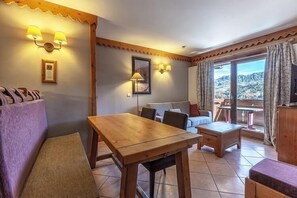 A cozy room with a wooden table, a sofa, and a view of a mountainous landscape through large windows. - 10m from ski slopes, 5th floor, view mountain, fitness, swimming pool, sauna, hammam, balcony, tv (La Plagne)