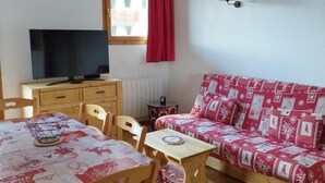 Una habitación acogedora con una mesa de madera, sillas, un sofá, un televisor y una ventana con cortina roja.