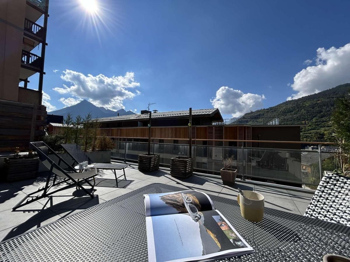 Une terrasse sur le toit, aménagée avec des chaises longues, une table et une balustrade en verre, offrant une vue panoramique sur un paysage montagneux.