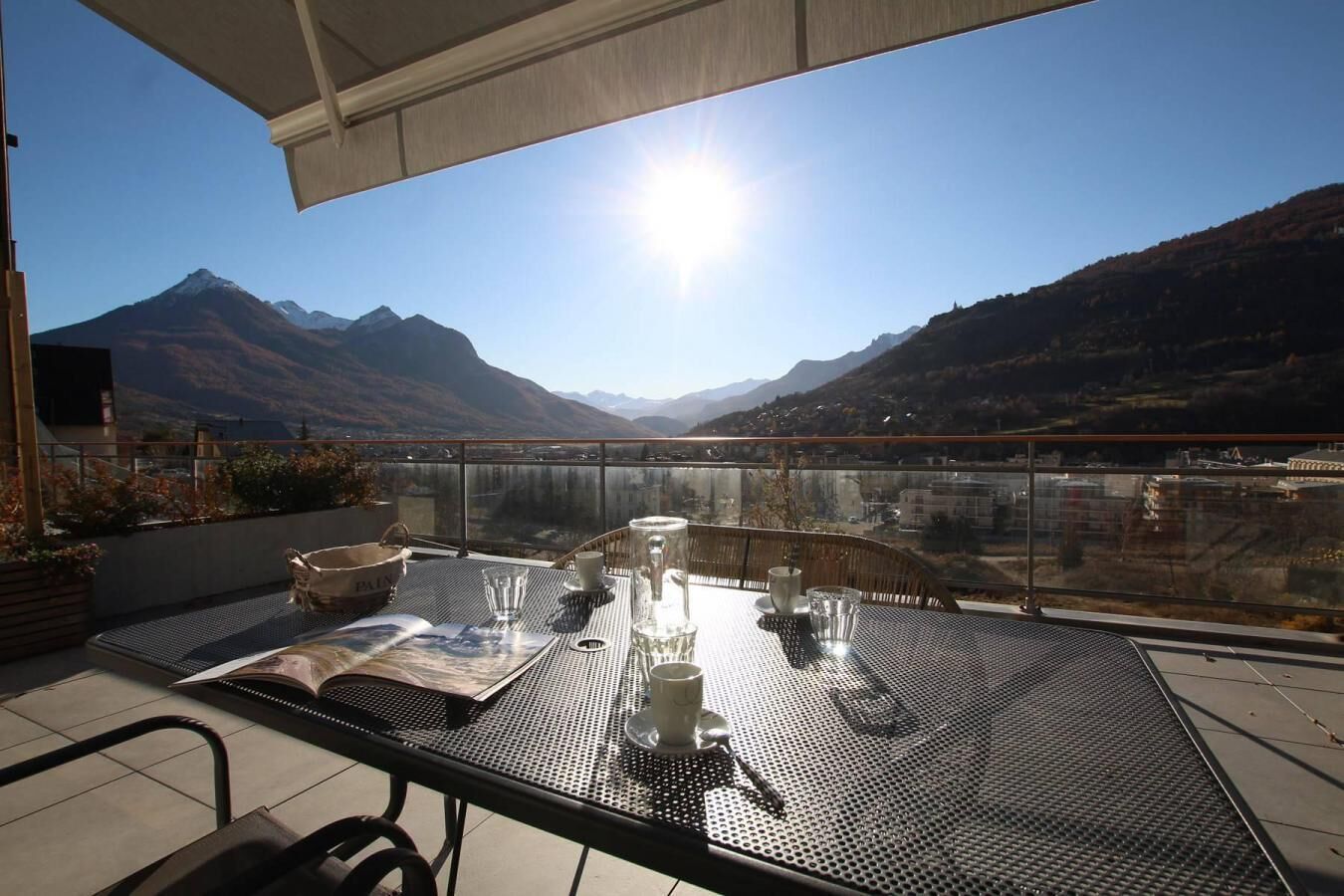 A balcony with a table set for dining, overlooking a mountainous landscape and a body of water.
