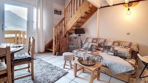 A cozy living room with a wooden staircase, a patterned sofa, a dining table with chairs, and a view of the outdoors.