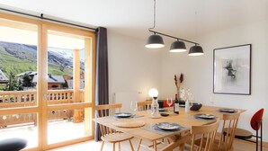 Un coin repas avec une table en bois dressée pour quatre personnes, une grande fenêtre offrant une vue sur un paysage montagneux et un luminaire suspendu moderne au-dessus de la table.