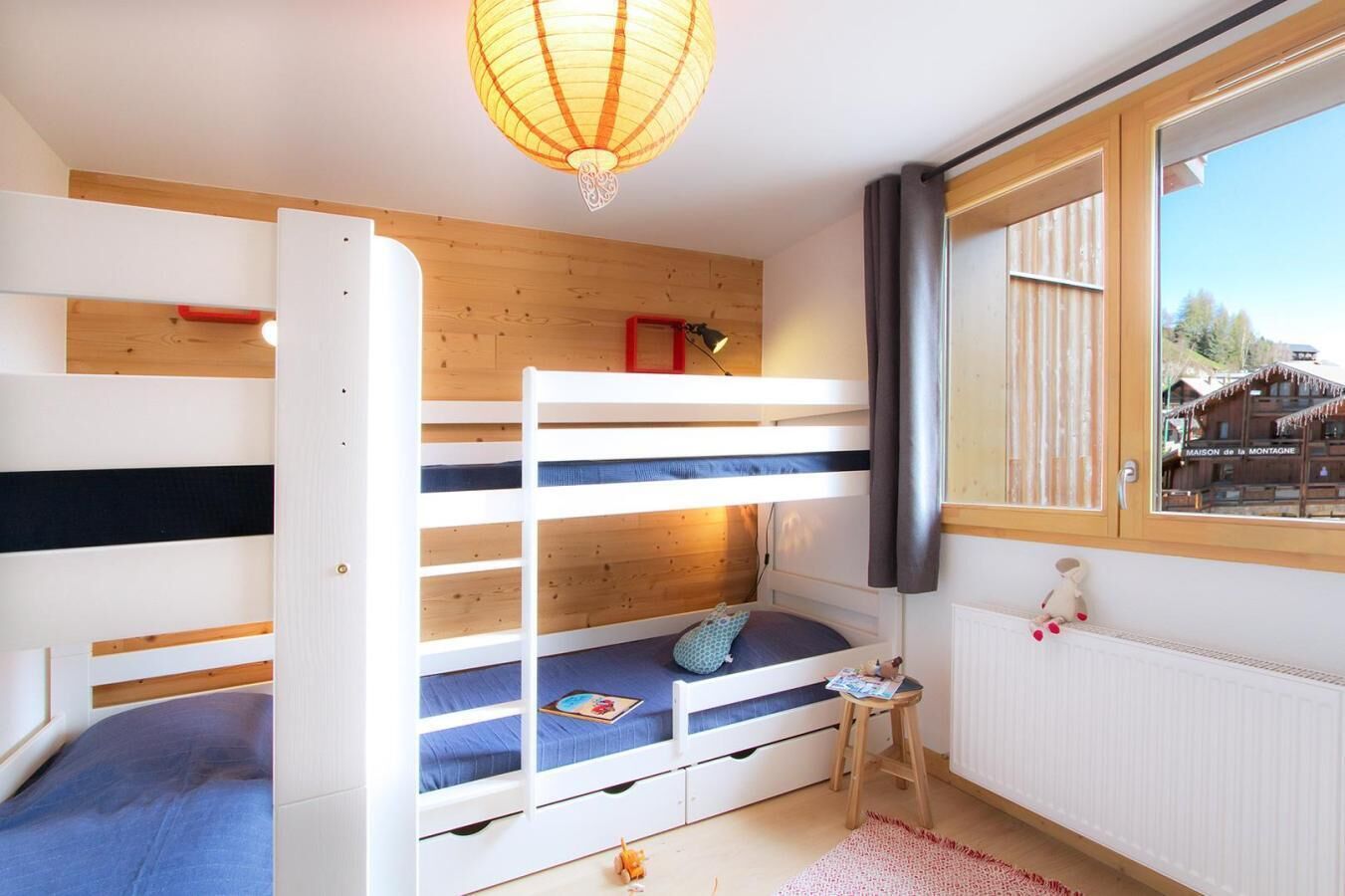 Ein Schlafzimmer mit Etagenbetten, Holzboden, einem kleinen Hocker und einem Fenster mit Blick auf ein Gebäude.
