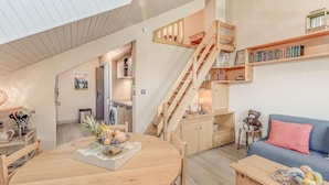 A cozy, well-lit room with a wooden dining table, a blue sofa, and a staircase leading to an upper level.