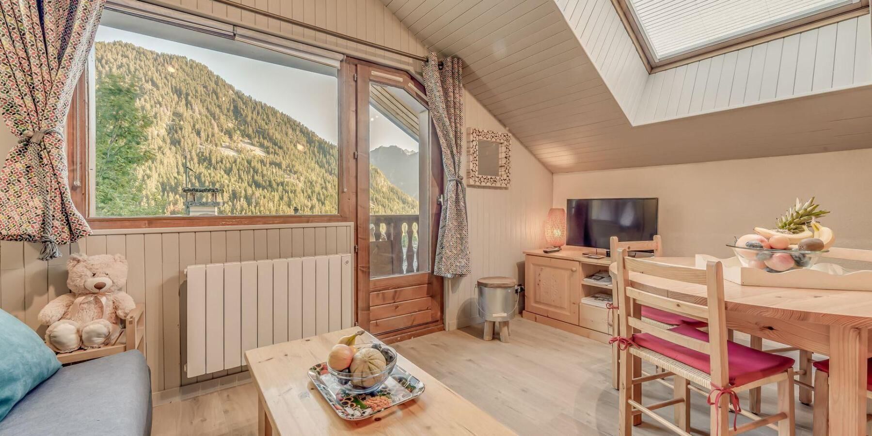 A cozy room with a large window, a wooden table with a fruit bowl, a TV, and a chair with a red cushion.
