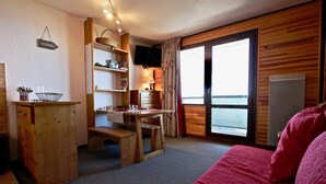 Una habitación acogedora con un escritorio de madera, un banco, una cama con una manta roja y un ventanal con cortinas.