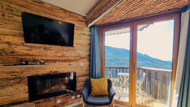 A room with a wooden wall, a fireplace, a TV, and a balcony with a view of the mountains.