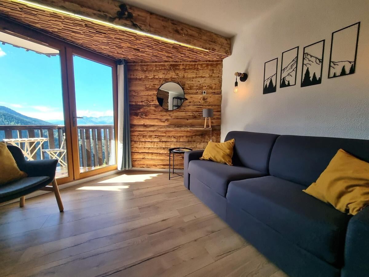 A living room with a sofa and armchair, wooden walls, a balcony with a view, and framed pictures on the wall.
