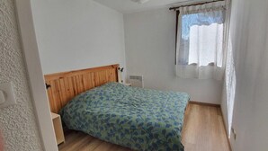 A small, simple bedroom with a wooden headboard, a single bed with a patterned bedspread, a nightstand, a window with a curtain, and a radiator. - Ground floor, terrace, parking, tv, ski locker, 65m², Serre Chevalier (La Salle-les-Alpes)