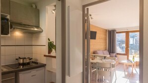 Eine kompakte Küche mit Herd, kleinem Kühlschrank und Mikrowelle. Angrenzend befindet sich ein Essbereich mit Tisch und Stühlen sowie eine Sitzecke. Das Zimmer verfügt über ein großes Fenster, das für natürliches Licht sorgt.