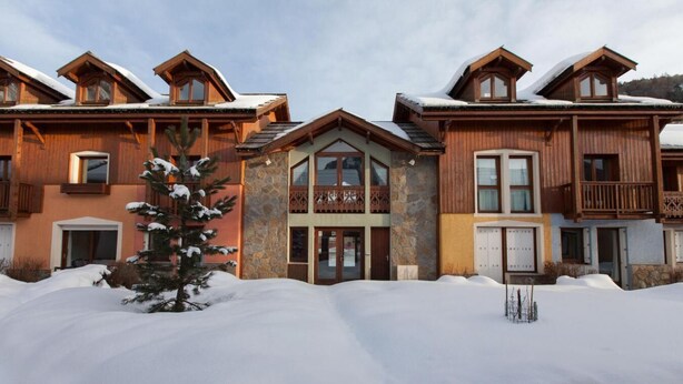 A row of wooden cabins with a stone facade, snow-covered roofs, and a central entrance with glass doors.