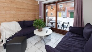 A cozy room with a wooden wall, a dark sofa, a small round table with a plant, and a view of a snowy outdoor area through a glass door. - Chalet, terrace, parking, tv, 61m², Serre Chevalier (La Salle-les-Alpes)
