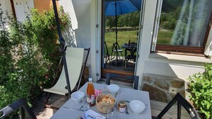 Un área de comedor al aire libre con una mesa para dos, bajo una sombrilla azul, con sillas y vistas a un jardín y un edificio.