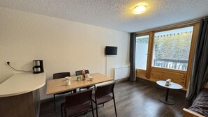 Quarto com mesa de jantar de madeira, quatro cadeiras, uma mesinha redonda com uma xícara, TV de tela plana na parede e janela com vista para árvores e uma varanda.