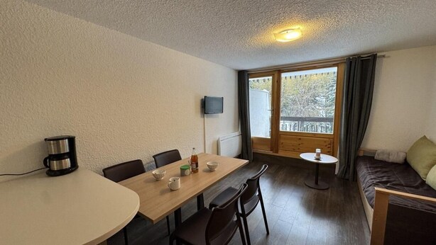 Um quarto acolhedor com uma mesa de jantar de madeira, cadeiras, uma pequena mesa redonda, um sofá e uma televisão presa à parede.