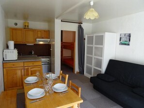 A compact kitchen with wooden cabinets, a microwave, and a stove. There is a dining table set with plates and glasses. A black sofa is in the living area, and a white cabinet is visible.
