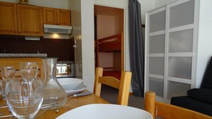 A compact kitchen with wooden cabinets, a dining table with white plates and glasses, and a bunk bed in the background. - 50m from resort center, 5th floor, balcony, tv, ski locker, 28m², Serre Chevalier (La Salle-les-Alpes)