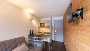Un espace de vie compact comprenant un coin cuisine, un téléviseur à écran plat fixé au mur et un canapé avec des coussins.