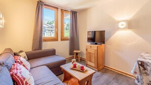 A cozy living room with a sofa, a small table with decorative items, a TV on a wooden cabinet, and a window with curtains.