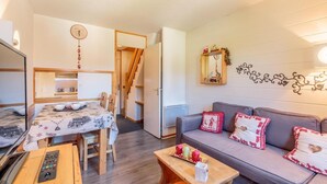 A cozy living room with a sofa, a dining table with chairs, a small kitchen area, and a staircase leading upstairs.