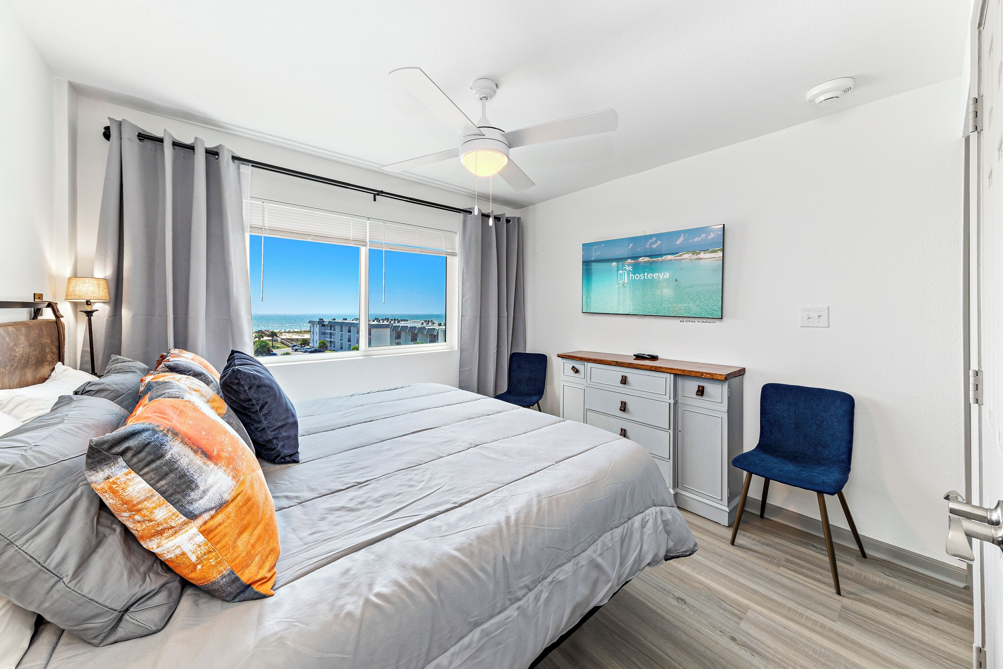 A bedroom with a large bed, a desk, a chair, a ceiling fan, and a window with a city view.