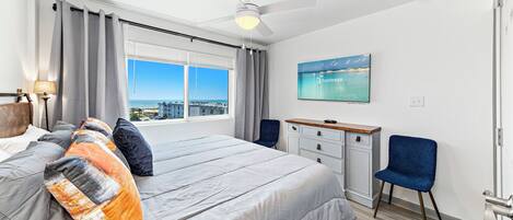 A bedroom with a large bed, a desk, a chair, a ceiling fan, and a window with a city view.
