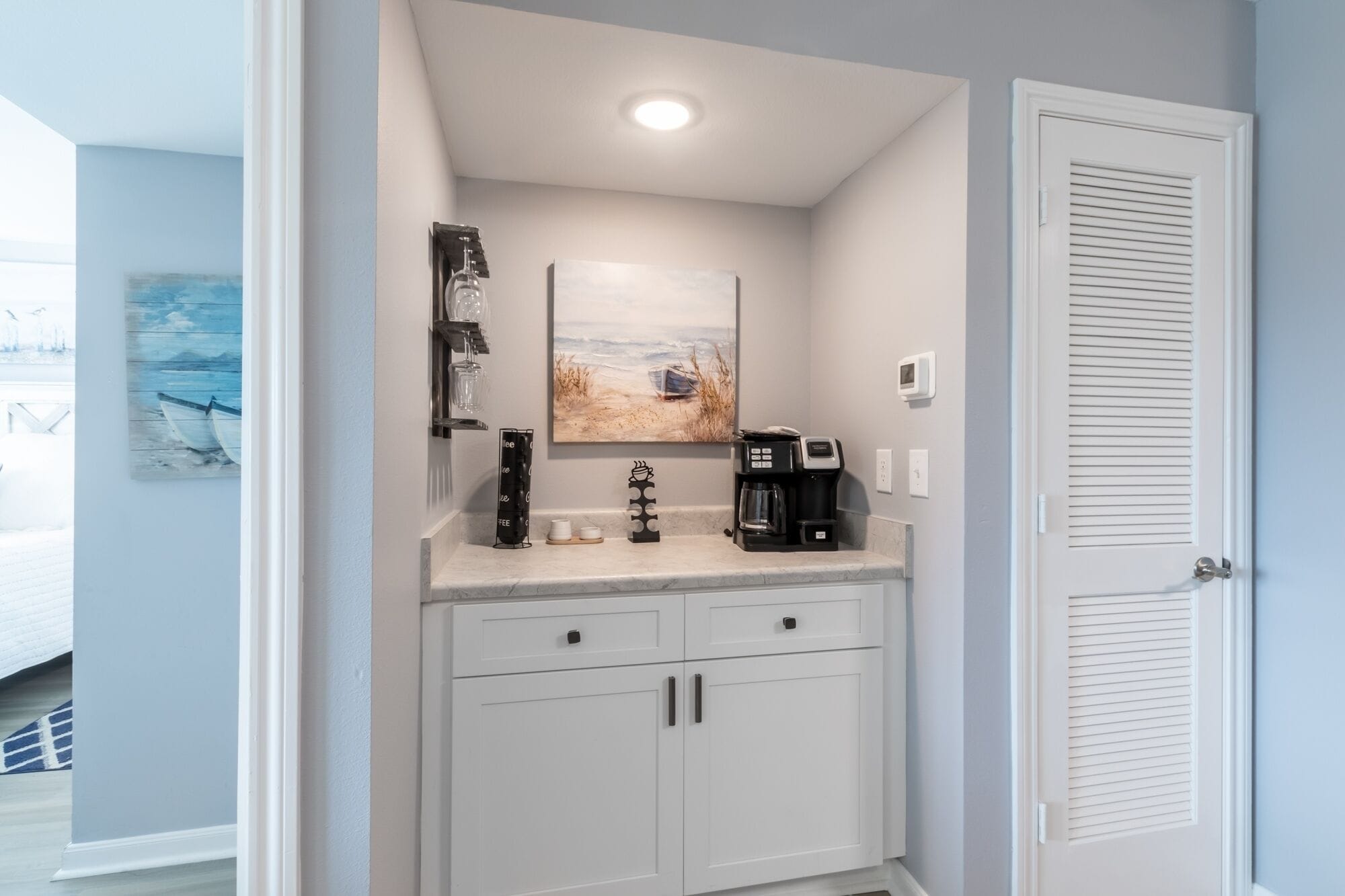 A compact kitchenette with a coffee maker, a wine glass holder, and a painting of a beach scene.