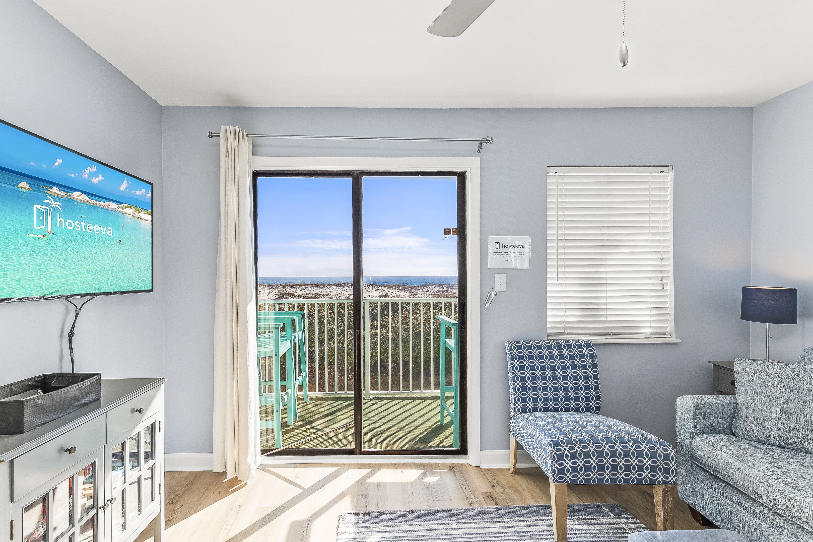 A modern living room with a large TV displaying a scenic view, a sliding glass door to a balcony, a patterned chair, and a grey sofa.