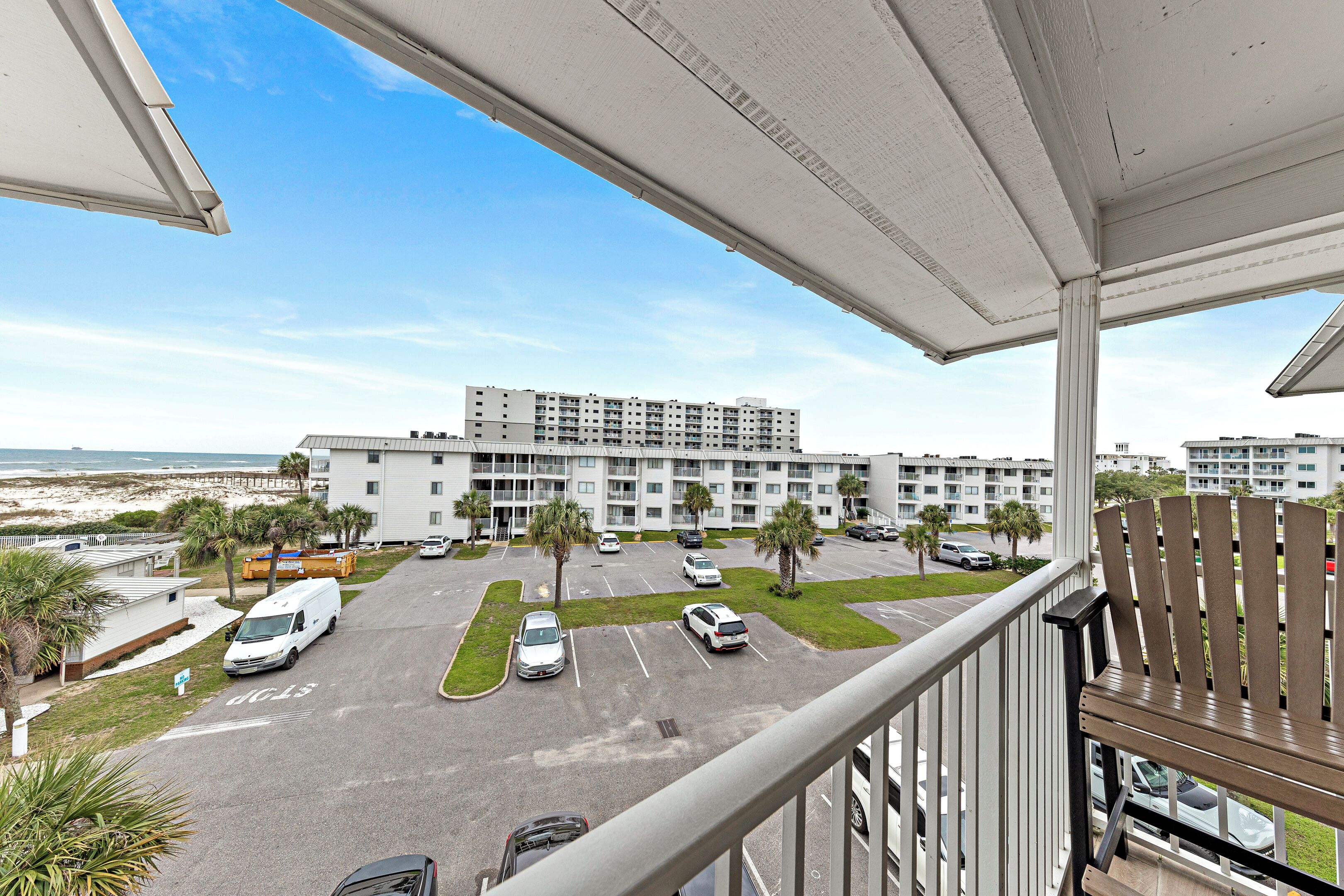 A view from a balcony overlooking a parking lot with several cars, a building with multiple floors, and a beach in the background.