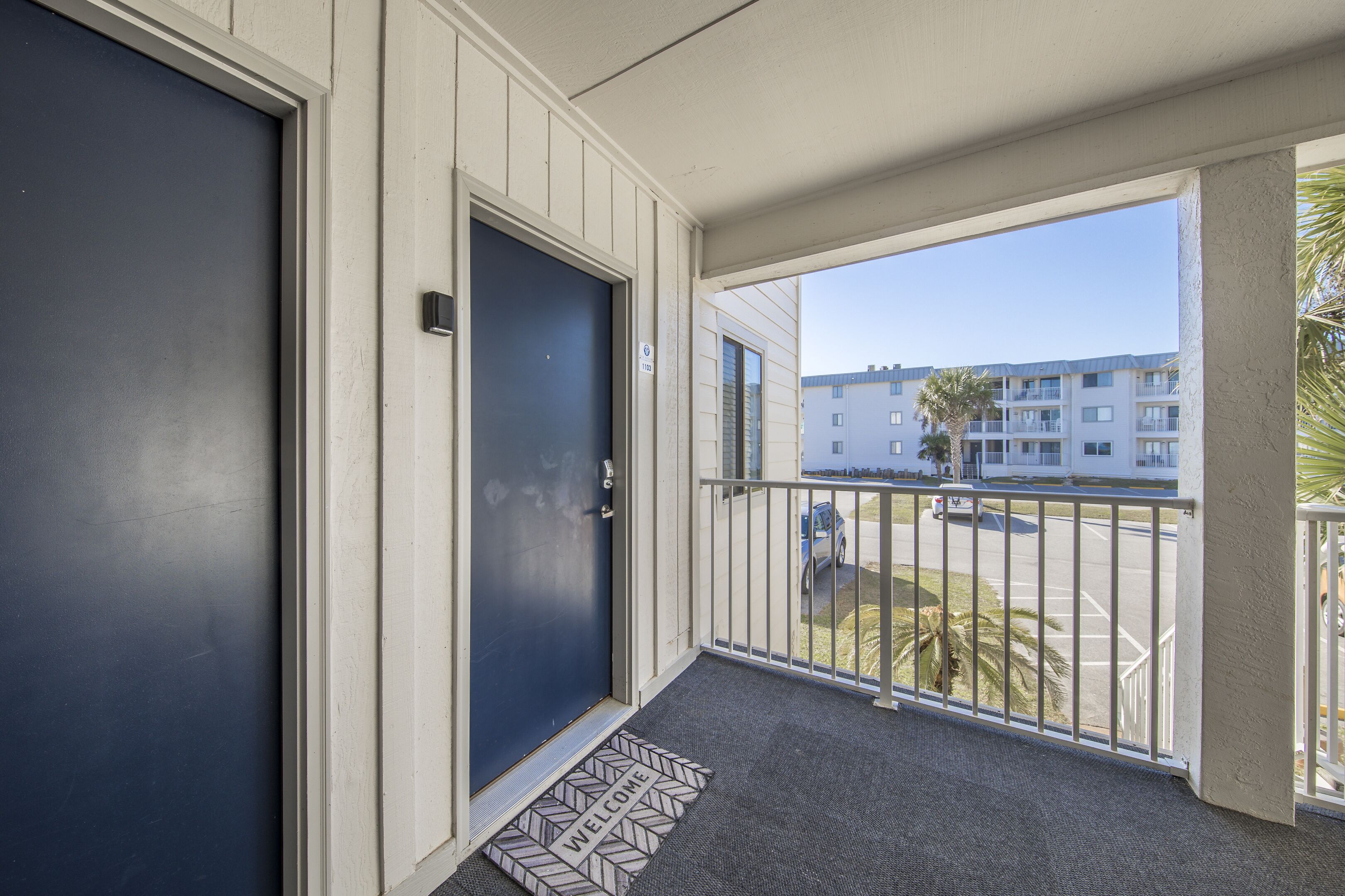 A hotel entrance with a dark blue door, a welcome mat, and a balcony with a railing.