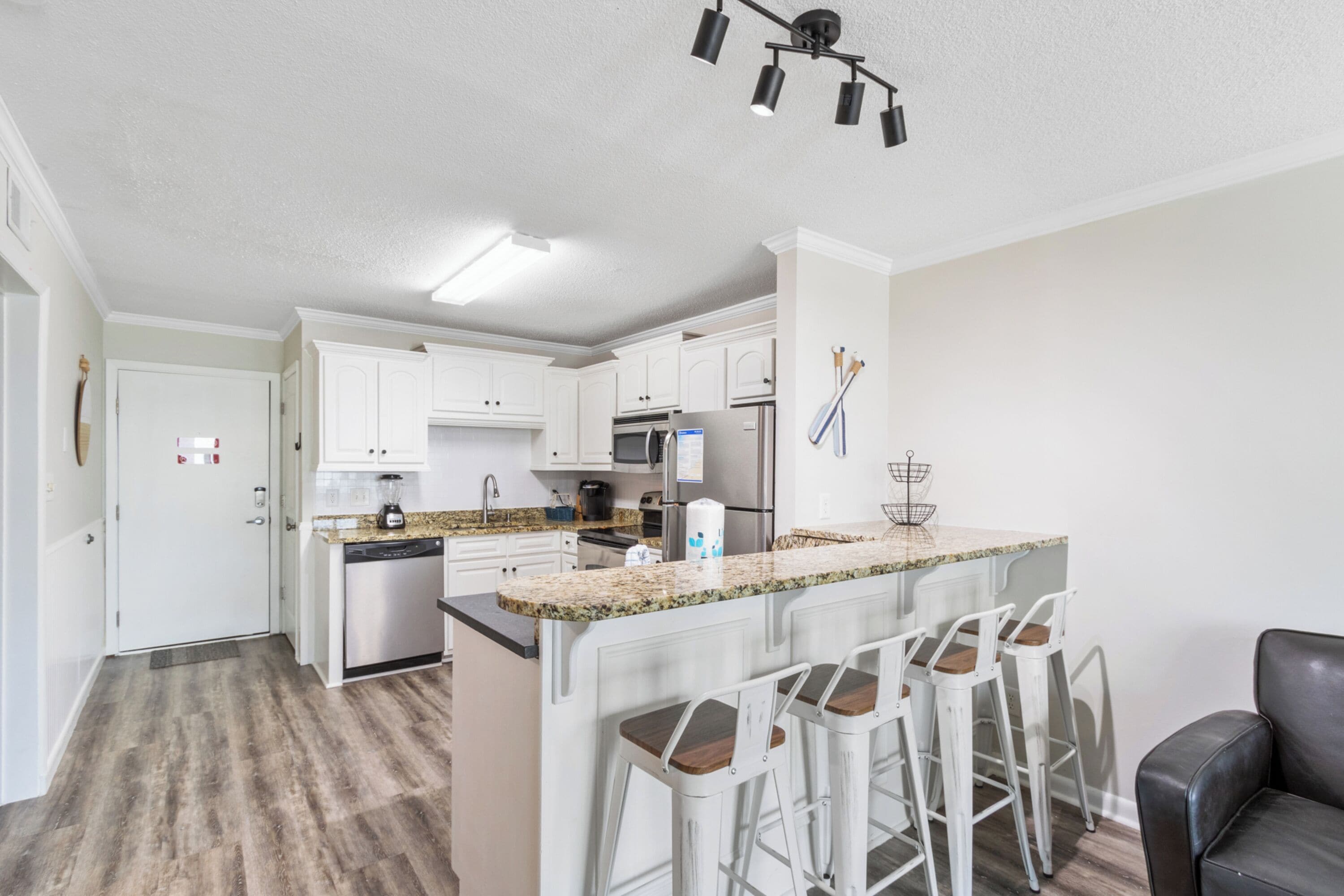A modern kitchen with white cabinets, a granite countertop, and stainless steel appliances. There are three bar stools and a black leather chair.