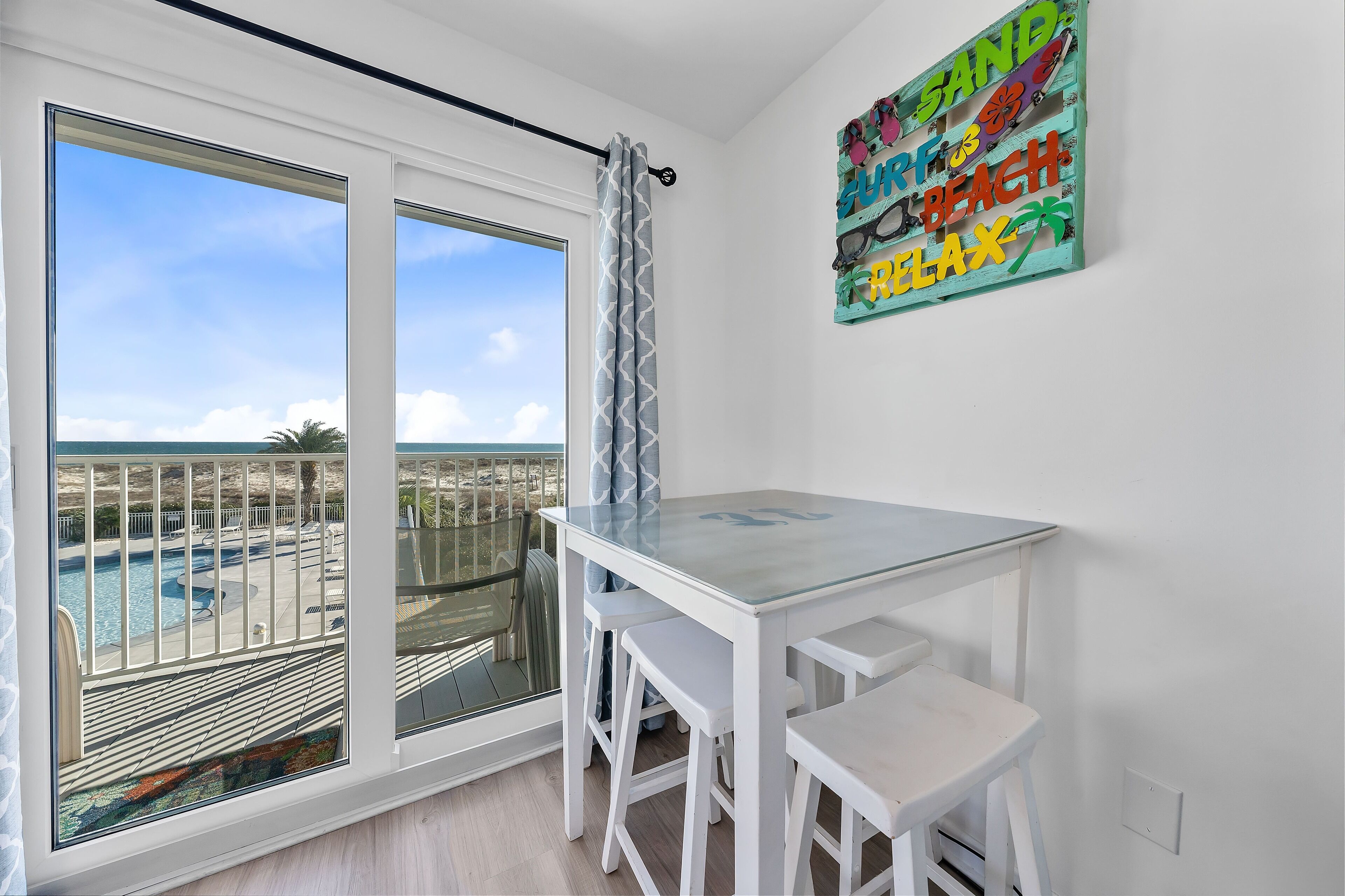 A balcony with a pool view, a glass table with white chairs, and a colorful artwork on the wall.