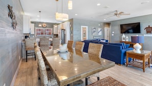 A modern living room with a large TV, blue sofas, and a dining area with wicker chairs. - Turtle Tracks | Outdoor Pool | Steps from Beach | Waterfront | My Beach Getaways (Gulf Shores)