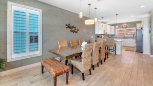 A modern kitchen with white cabinets, a stainless steel refrigerator, and a dining area with wicker chairs and a glass table. - Turtle Tracks | Outdoor Pool | Steps from Beach | Waterfront | My Beach Getaways (Gulf Shores)