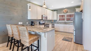 A kitchen with white cabinets, a marble countertop, a stainless steel refrigerator, and a bar area with wicker chairs. - Turtle Tracks | Outdoor Pool | Steps from Beach | Waterfront | My Beach Getaways (Gulf Shores)