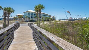 A wooden boardwalk leading to a beachfront property with multiple-story buildings, palm trees, and a clear blue sky. - Turtle Tracks | Outdoor Pool | Steps from Beach | Waterfront | My Beach Getaways (Gulf Shores)